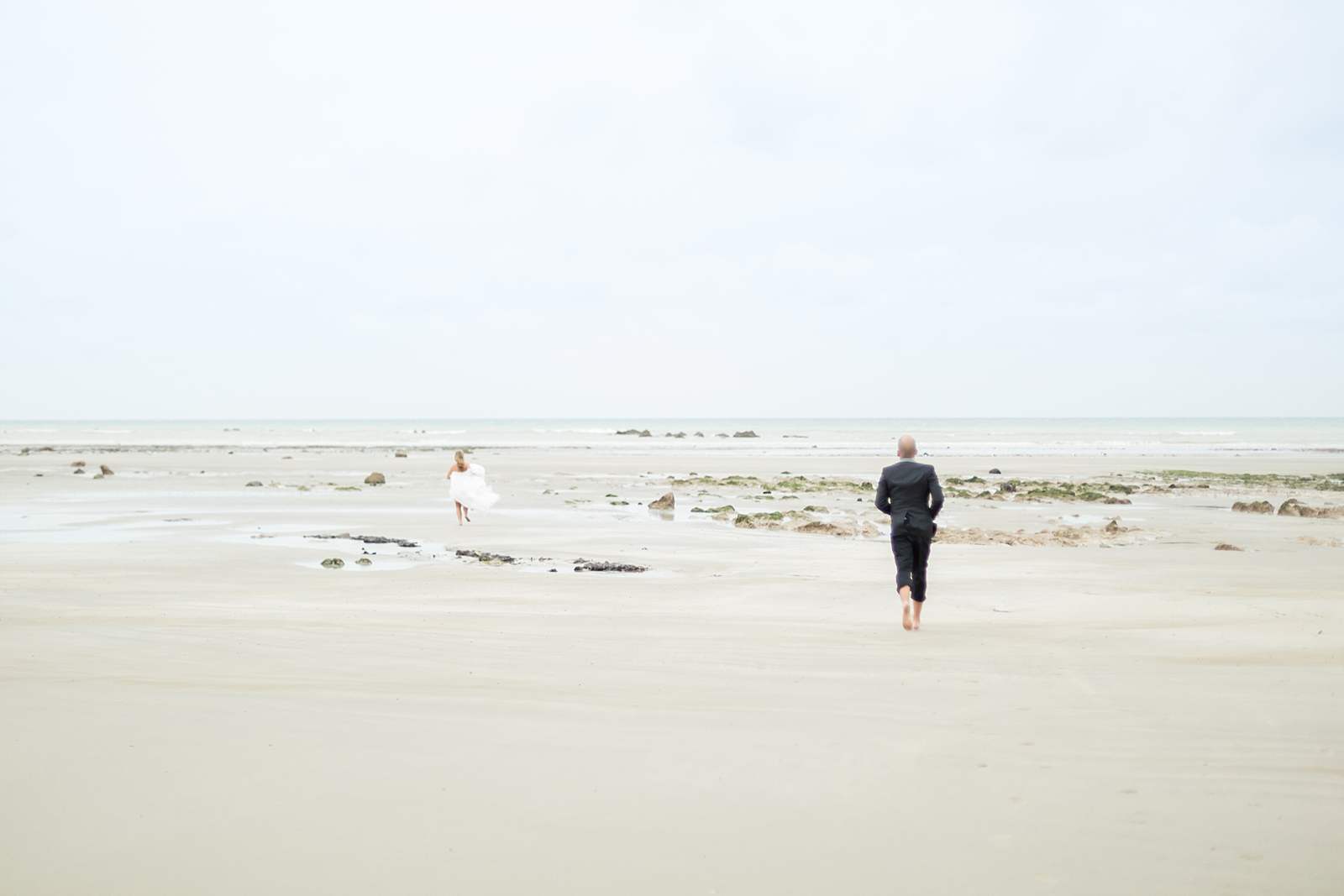 les mariés se courent après sur la plage de normandie. ils sont pieds nus dans le sable. la marée est basse.
